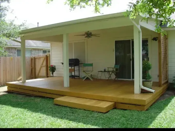 Small wooden backyard deck with a covered roof, ceiling fan, chairs, and a grill beside a fenced lawn.
