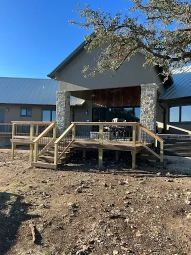 Wooden porch with stone columns and railing on a house under construction, surrounded by bare dirt.