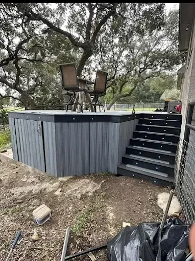 Raised outdoor deck with stairs, surrounded by trees and dirt, with two chairs on top