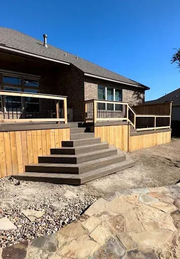 Stairway to a raised wooden deck on a brick house, with stone patio and clear blue sky.
