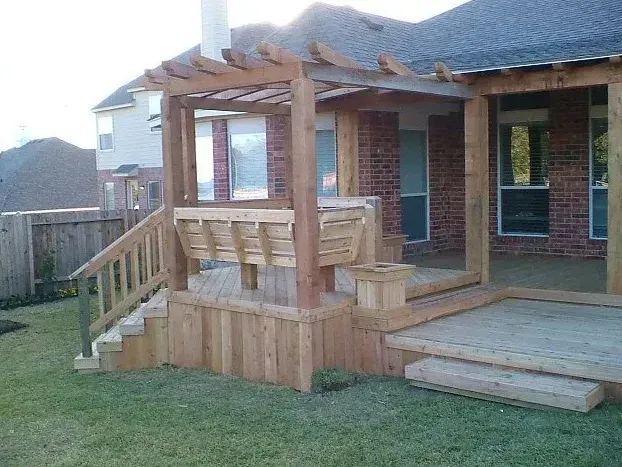 Wooden front porch with steps and railing on a brick house, beside a lawn and fenced yard.