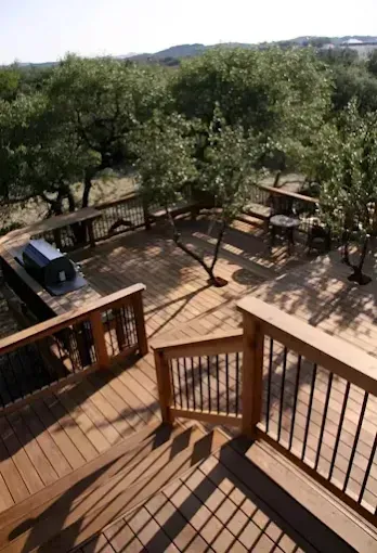 Wooden deck overlooking leafy trees, with railings, stairs, and a parked vehicle in the shade.