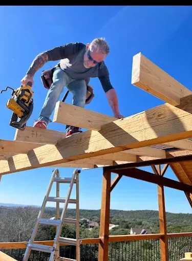 Worker kneels on roof beams using a yellow power saw under a clear blue sky