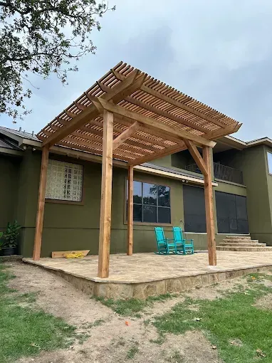 Wooden pergola over a patio beside a house, with two turquoise chairs and steps leading to the yard.