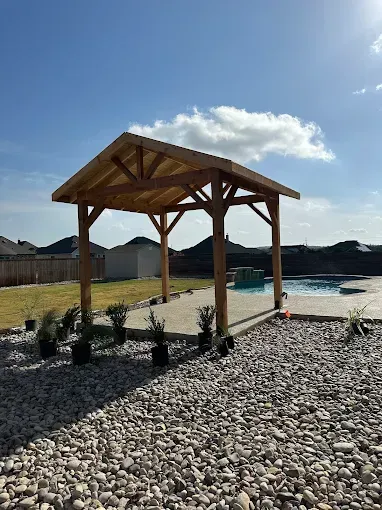 Wooden pavilion on a gravel lot under a bright blue sky with scattered clouds.