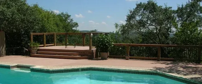 Outdoor pool with a wooden deck and railing overlooking trees under a blue sky
