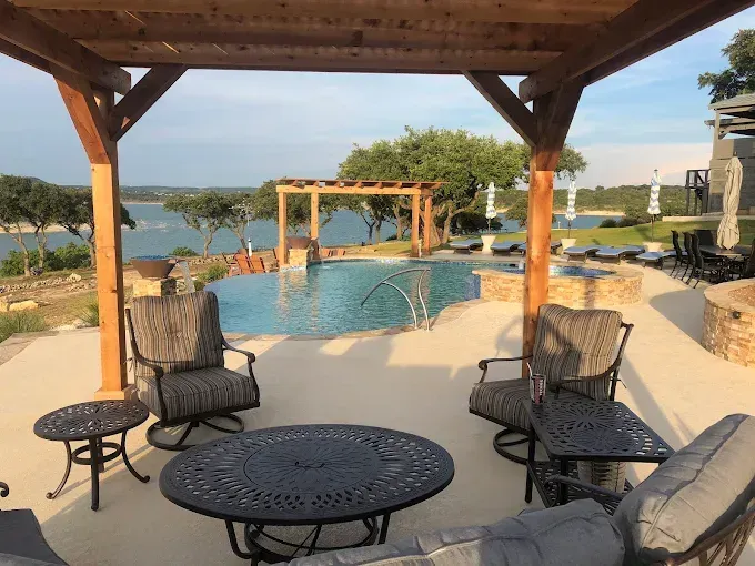 Poolside patio with wicker chairs and table under a wooden pergola, overlooking a waterfront view