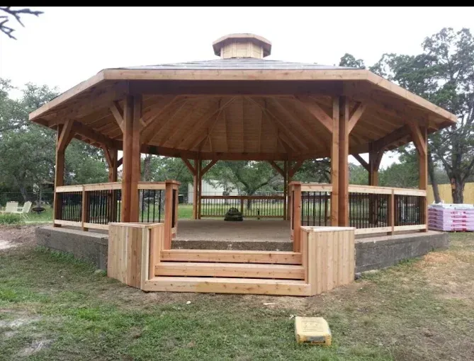 Wooden octagonal gazebo with steps in a grassy park, with trees and benches in the background