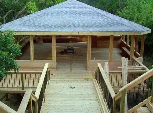 Wooden walkway leading to a pavilion with a gray roof, surrounded by greenery.