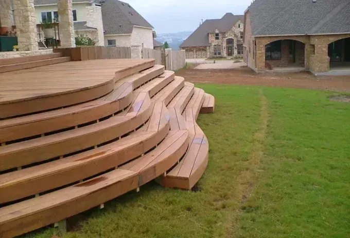 Curved wooden tiered seating beside a grassy lawn in a suburban backyard.