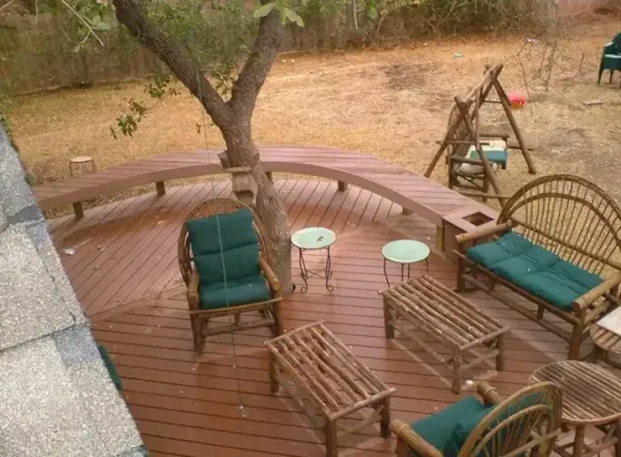 Wooden deck patio with green cushioned chairs, tables, a bench, and a tree overlooking a dry yard.