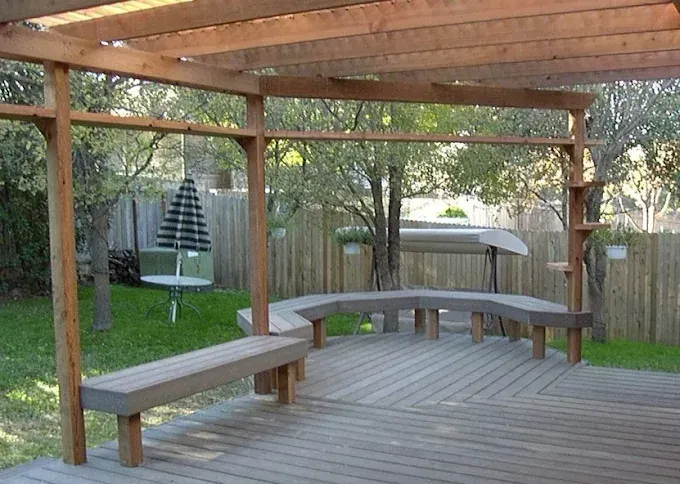 Covered wooden deck with built-in benches and a pergola in a backyard, bordered by trees and a fence
