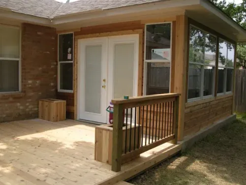 Wooden porch with white double doors, railing, brick siding, and large windows on a house