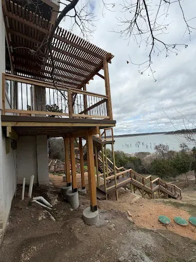 House under construction on a hillside with wooden deck, stairs, and lake view in the background