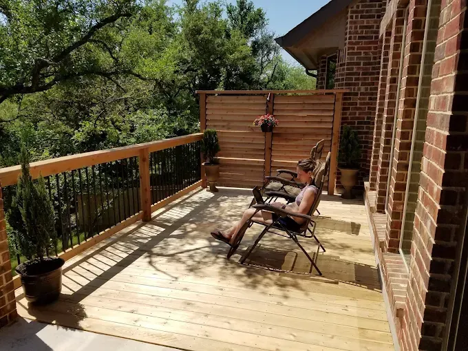 Wooden deck with lounge chairs beside a brick house, overlooking trees and a railing