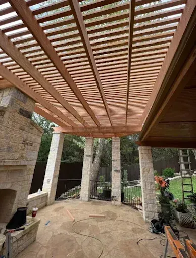 Covered patio with wooden slatted pergola, stone columns, and tiled floor beside a garden
