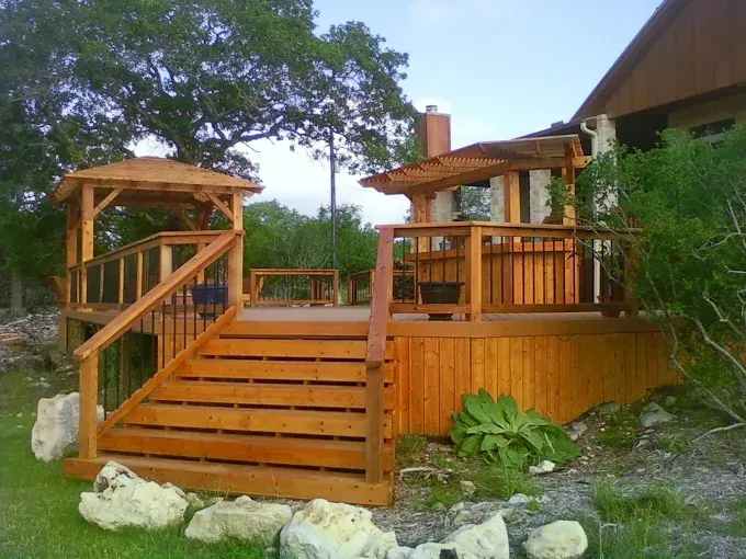 Wooden deck with stairs and railings on a landscaped hillside, with small pergola roofs and greenery around it