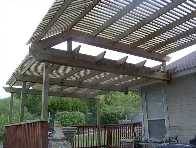 Covered wooden patio with slatted roof, railing, and outdoor seating beside a house and trees.