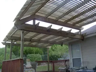 Covered wooden patio with slatted roof, railing, and outdoor seating beside a house and trees.
