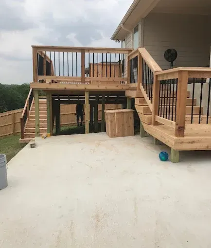 Wooden deck with railings and stairs beside a house, overlooking a concrete patio.