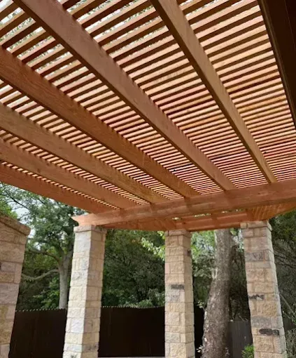 Wooden pergola with slatted roof supported by stone columns in a garden setting