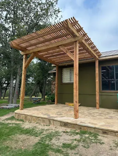 Wooden patio pergola beside a green house, with open slatted roof on a concrete porch.