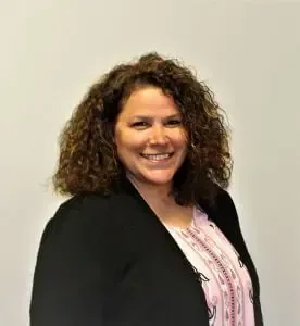 A person with curly brown hair wearing a black blazer over a light patterned top, smiling against a neutral background.