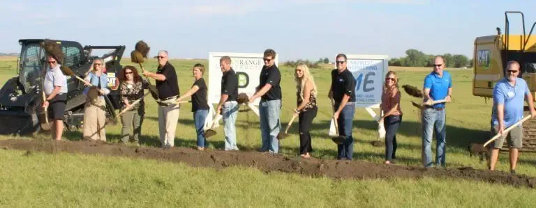 A group of people holding shovels perform a ceremonial groundbreaking in a field, with construction equipment nearby.