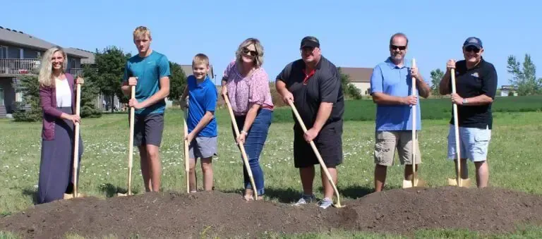 Seven people stand in a line on a grassy field, each holding a shovel in a ceremonial groundbreaking pose.