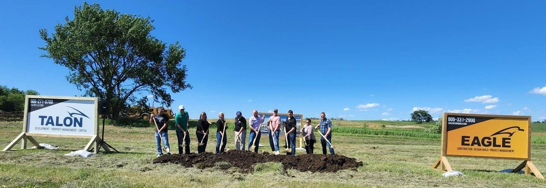 A group stands in a field for a ceremonial groundbreaking with shovels, flanked by Talon and Eagle branded signs.