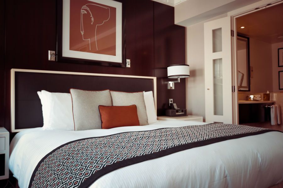 A modern hotel room with a neatly made bed, white linens, an orange accent pillow, and a minimalist abstract art print.
