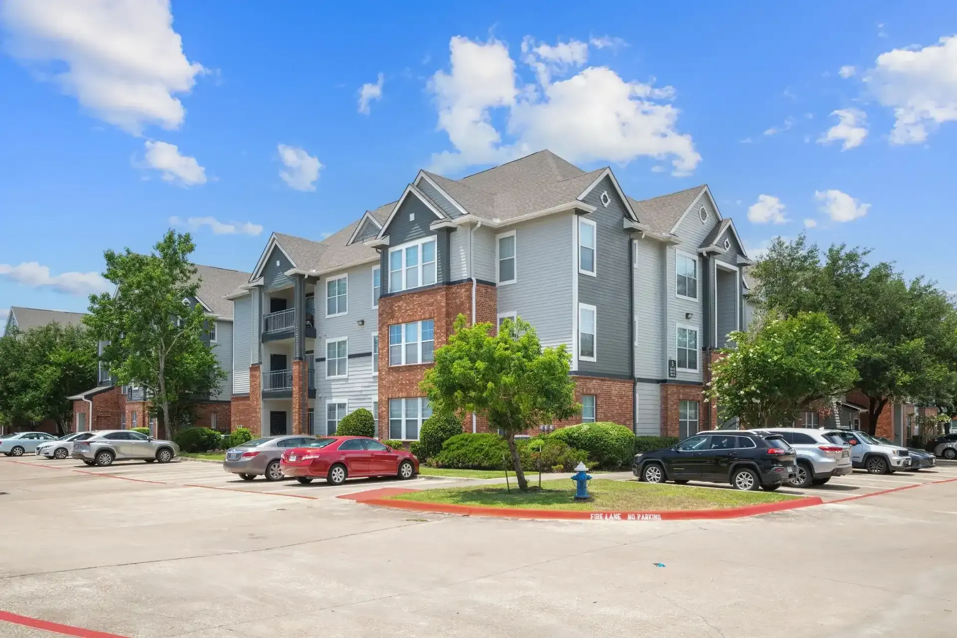 Apartment building with gray siding, brick accents, and parked cars on a sunny day.