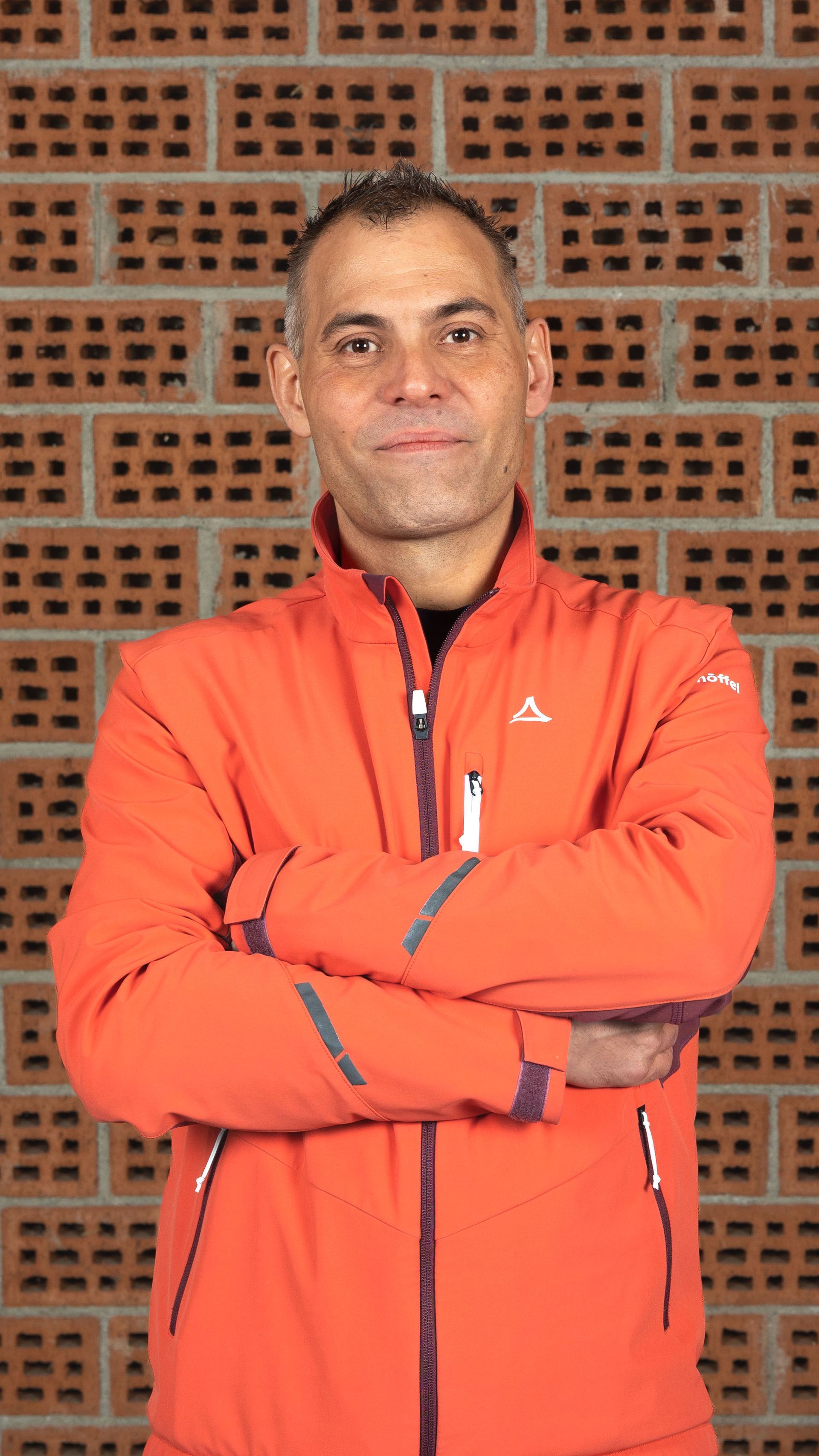 Eine Person in einer orangefarbenen Jacke steht mit verschränkten Armen an einer Backsteinmauer.