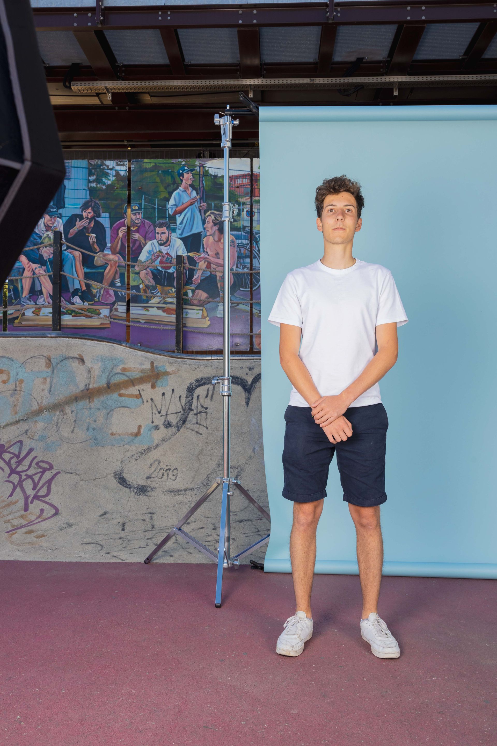 Ein junger Mann in einem weißen T-Shirt und Shorts steht vor einem blauen Hintergrund. Ein Blitzlicht ist am Rande des Bildes zusehen.