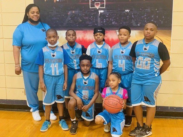 Youth basketball team in blue uniforms poses with coach in front of a basketball court mural.