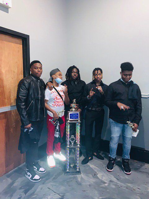 Five young people posing with a trophy. One wears a mask, others in black outfits, looking proud. Interior setting.