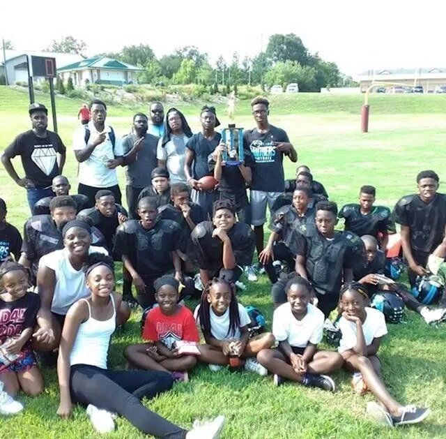 Youth football team poses outdoors with trophy. Players and coaches are mostly black.