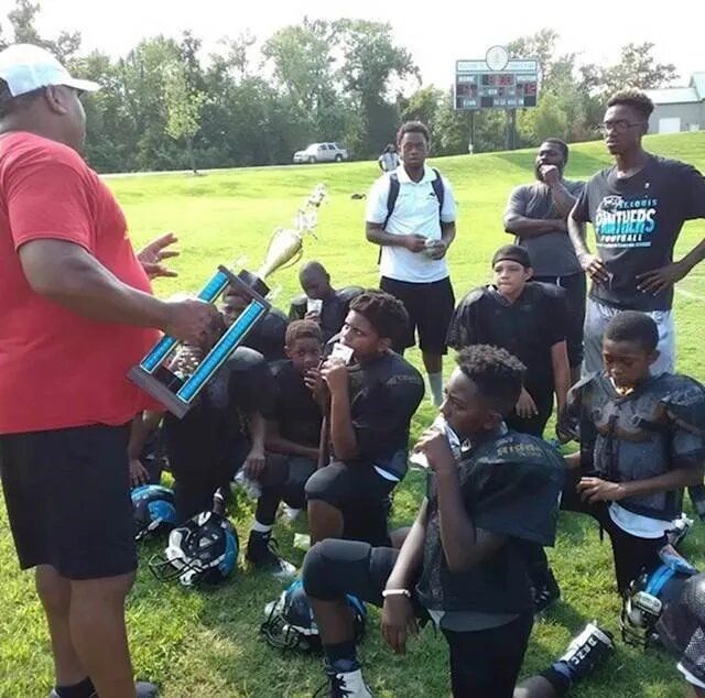 Coach holding trophy, surrounded by kneeling youth football players in black uniforms on a grassy field.