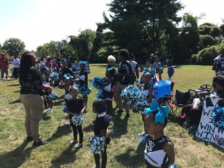 Youth cheerleaders in black and teal uniforms on a grassy field, with a group of adults.