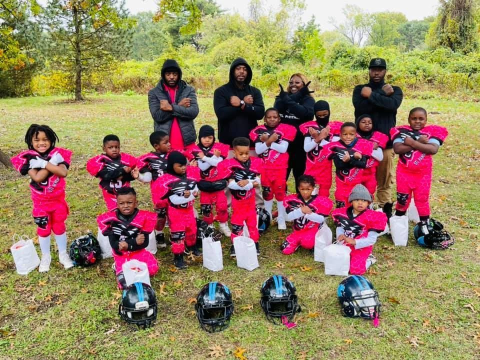 Youth football team in pink and red uniforms with coaches, outdoors. They make a crossed-arms gesture.