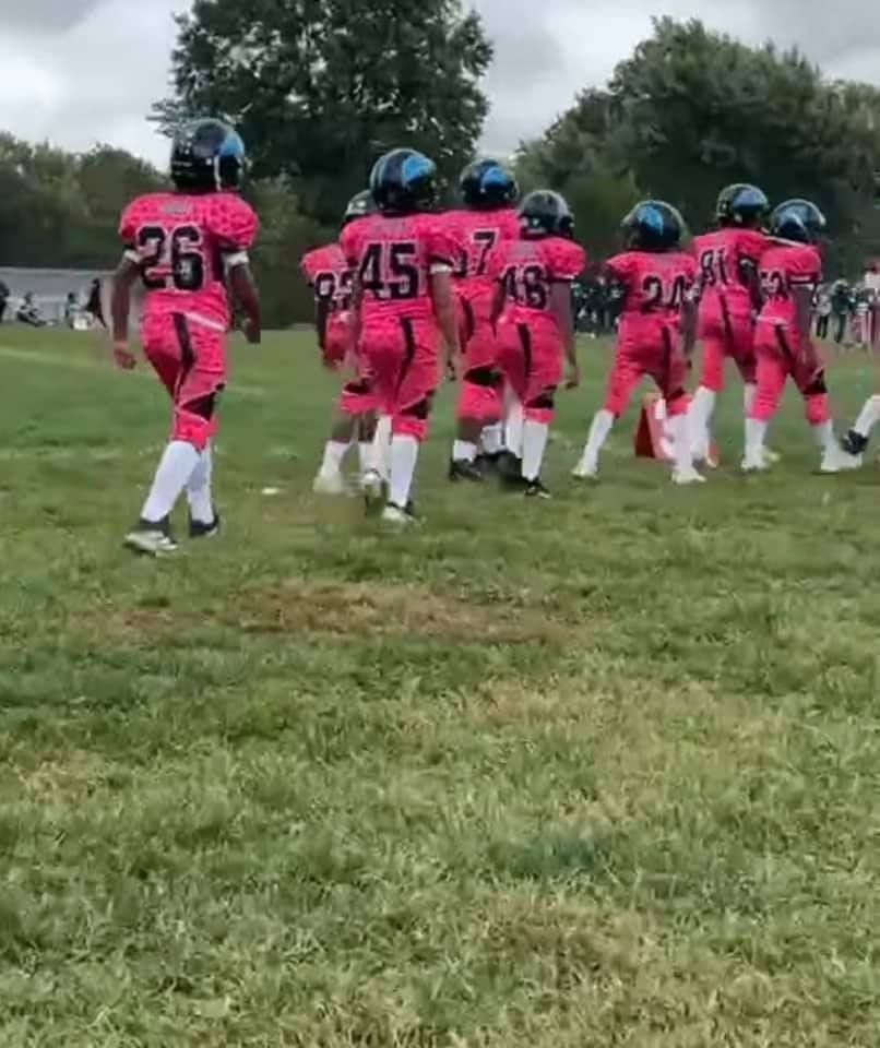 Youth football team in pink uniforms with black helmets on a grassy field.