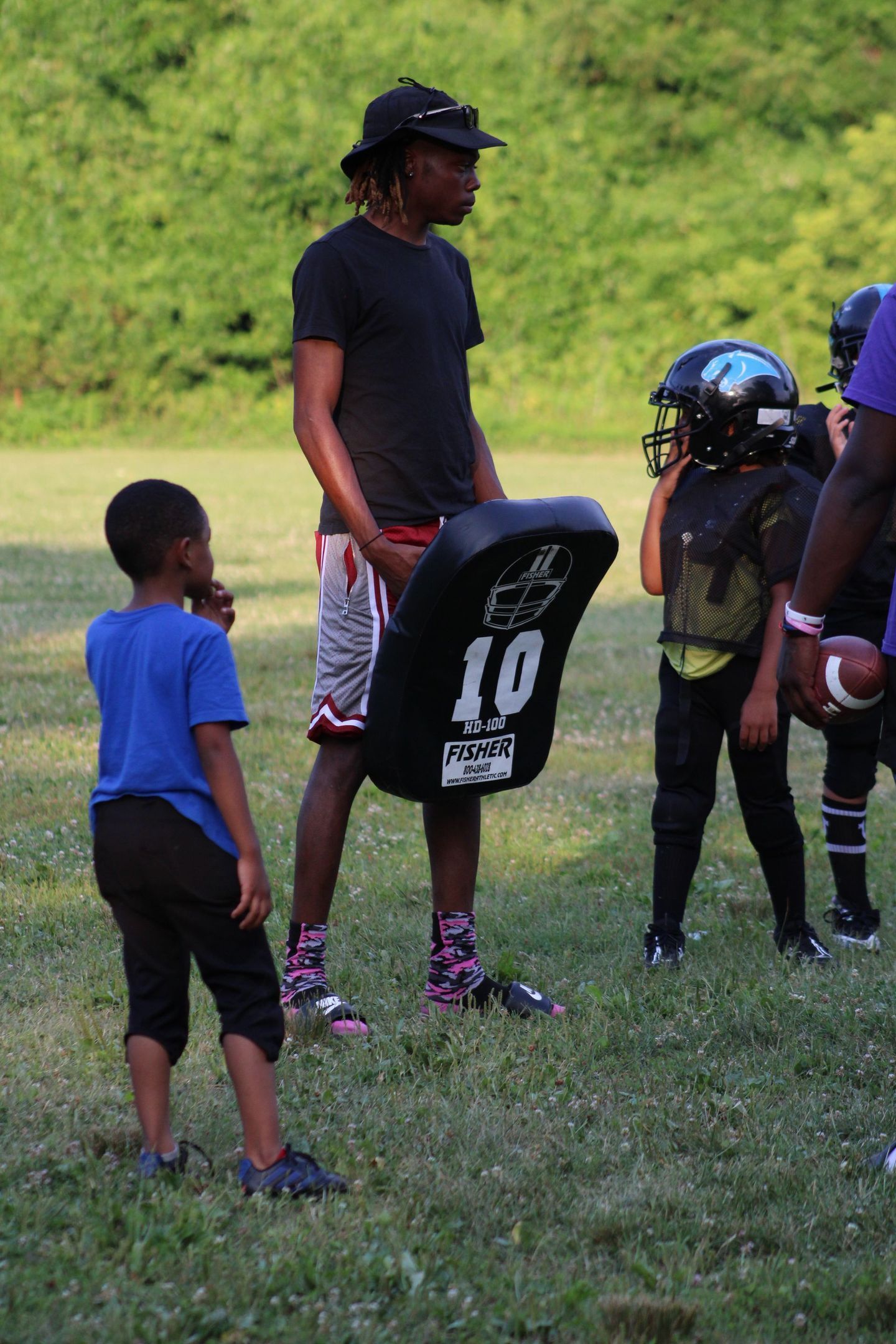A football coach holding a pad stands with young players on a grassy field.