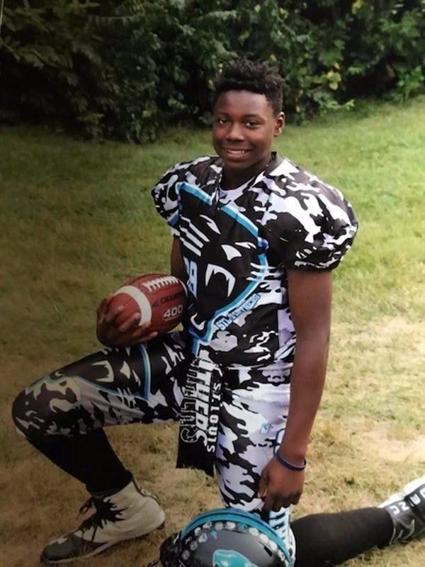 Football player kneels in jersey and pants with a black, white, and turquoise pattern, holding a ball.