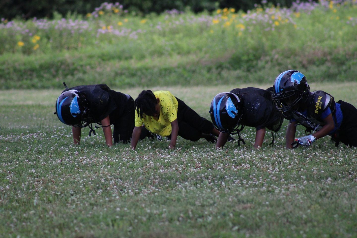 Football players in a line, doing push-ups on a grassy field. Three wear black and blue, one in yellow.