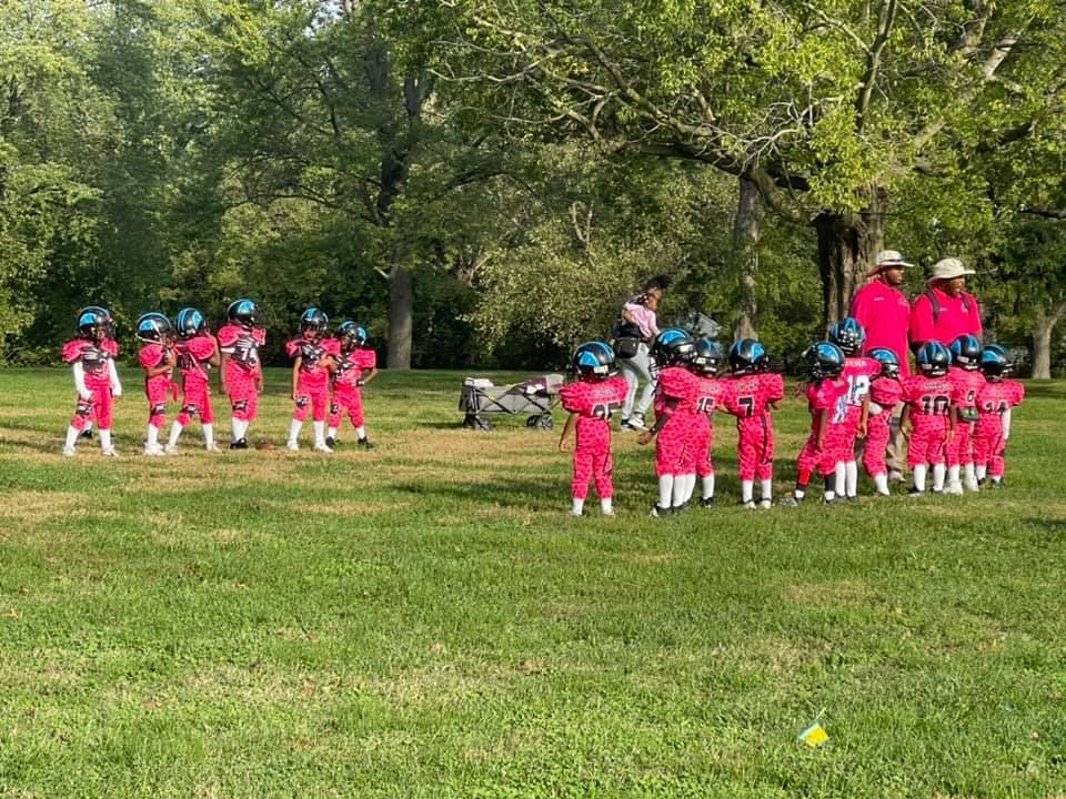 Youth football team in pink uniforms and helmets on a grassy field with coaches.