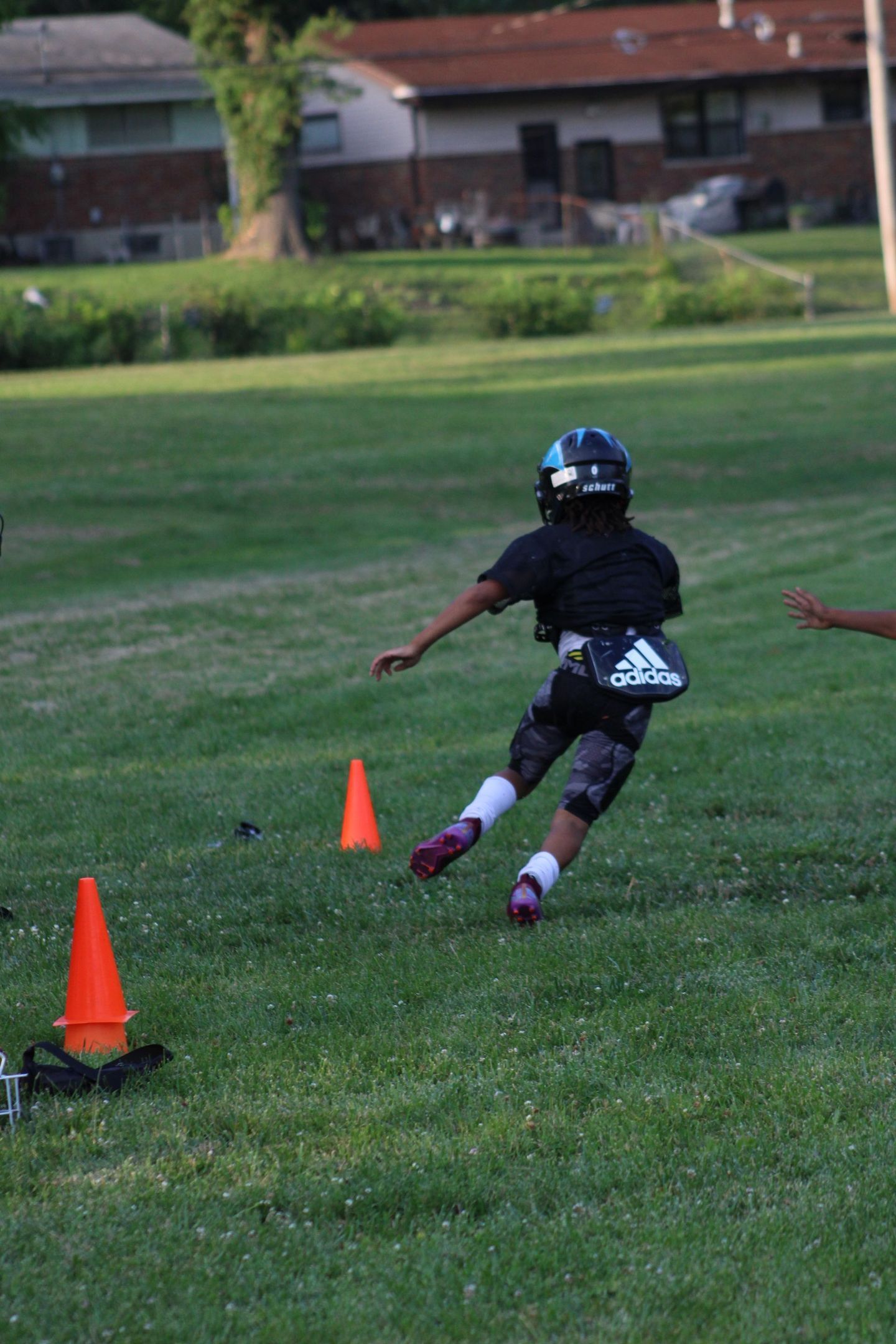 A young football player running on a grassy field, approaching cones, wearing a helmet and Adidas attire.