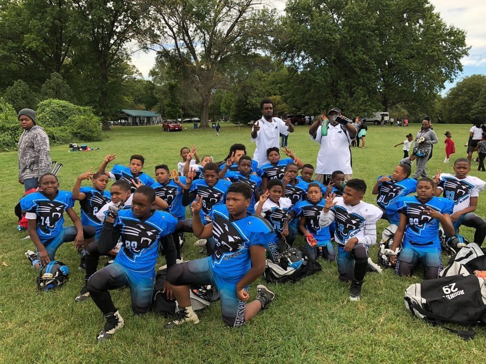 Youth football team in blue uniforms kneels on a grassy field. Coaches stand behind.