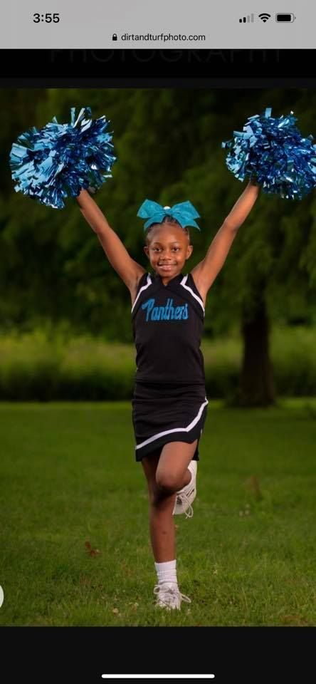 Young cheerleader with blue pom-poms, black uniform, and blue bow, smiles while posing outdoors.