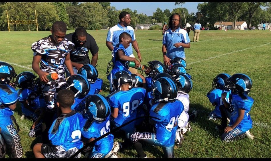 Youth football team huddle on a sunny field, wearing blue and black uniforms; coaches nearby.