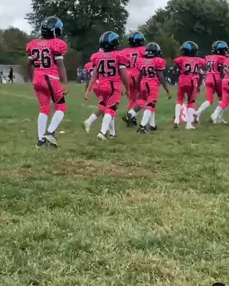 Youth football team in pink uniforms jogs on a grassy field. Players wear black helmets with blue visors.